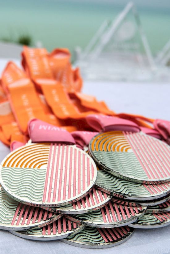 A stack of colorful medals with orange ribbons is arranged on a table, with a blurred outdoor background on a sunny day.