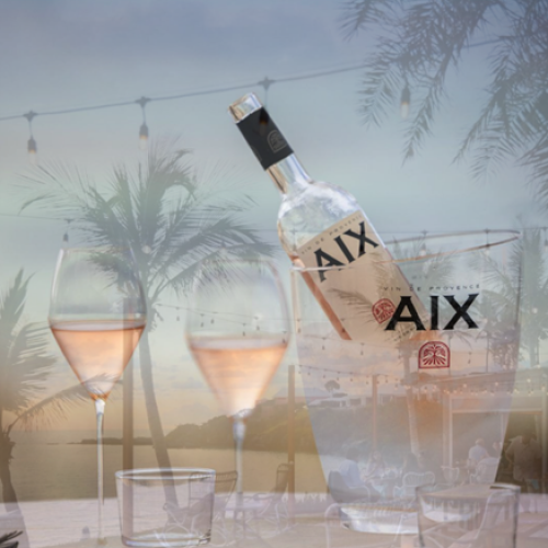 A bottle of AIX wine is in an ice bucket, surrounded by three filled wine glasses on an outdoor table with palm trees in the background and string lights.