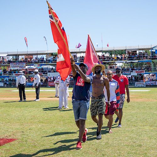 People walk on a grass field holding flags, with spectators in the background at a sporting event.