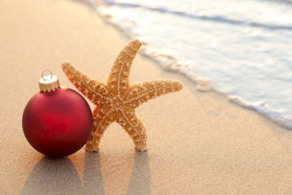 A red Christmas ornament and a starfish are placed on a sandy beach with gentle waves in the background.
