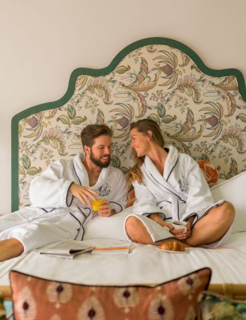 A couple is sitting on a bed in bathrobes, enjoying drinks and having a relaxed conversation in a cozy, well-decorated bedroom with patterned headboard.