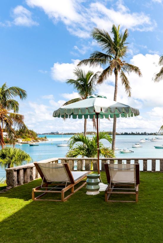 A seaside scene with palm trees, lounge chairs, an umbrella, and a pink building overlooking a bright blue ocean with distant boats and clear skies.