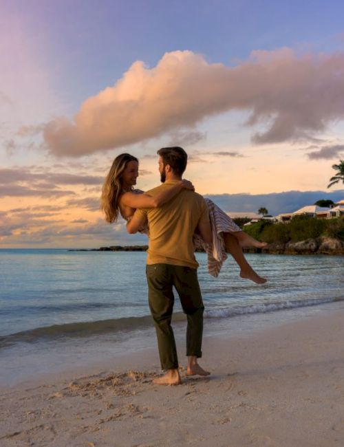 A couple is at the beach during sunset, with the man lifting the woman in his arms. The sky is colorful, and there are palm trees and homes in the background.