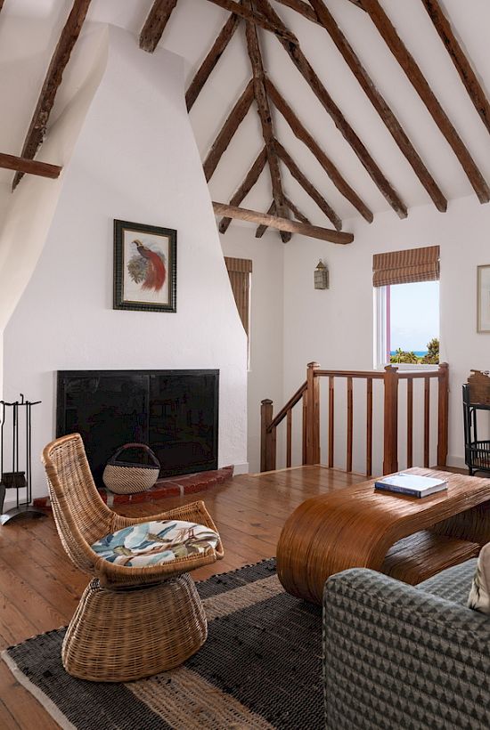 A cozy living room with wooden beams, a fireplace, wicker chair, sofa, coffee table, and desk. Windows provide a scenic ocean view.