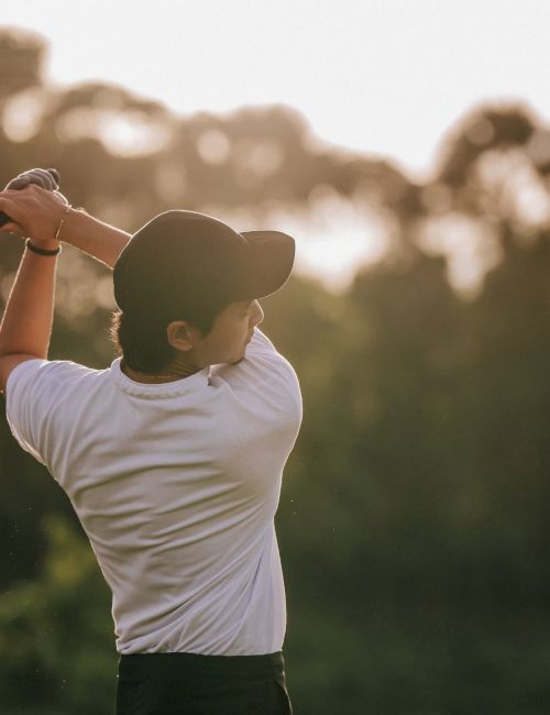 A person wearing a white shirt and black cap is swinging a golf club on a golf course with a blurry background of trees and sunlight.