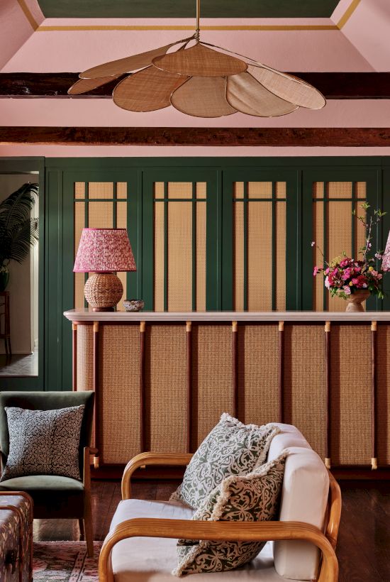 A cozy living room with pink walls, green accents, wooden furniture, floral lampshades, and a leafy ceiling fan. Fresh flowers add charm to the space.