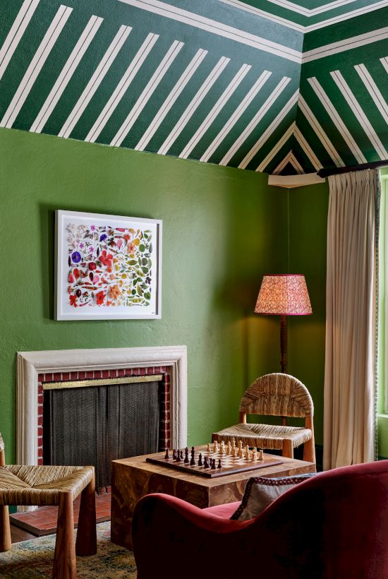 A room with green walls, a striped ceiling, a fireplace, a chessboard on a table, wicker chairs, a window with curtains, and a colorful framed picture.