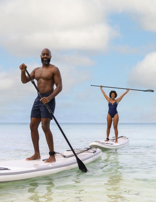 Two people are paddleboarding on clear, calm water under a bright sky; one stands confidently while the other raises a paddle in the air.