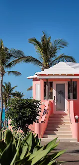 Pink seaside villa with white shutters, lush palm trees, sunny blue sky, and turquoise ocean below.