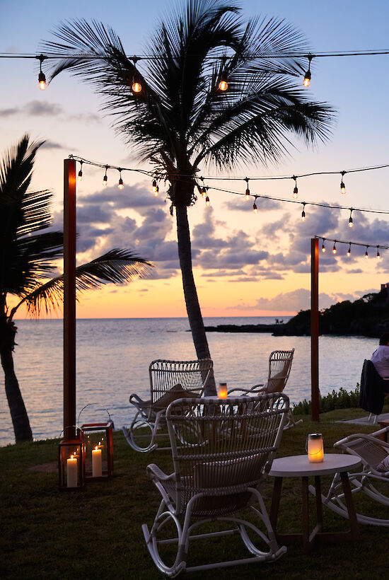 An outdoor restaurant or bar by the beach at sunset, decorated with string lights and palm trees, with patrons dining under a covered area.