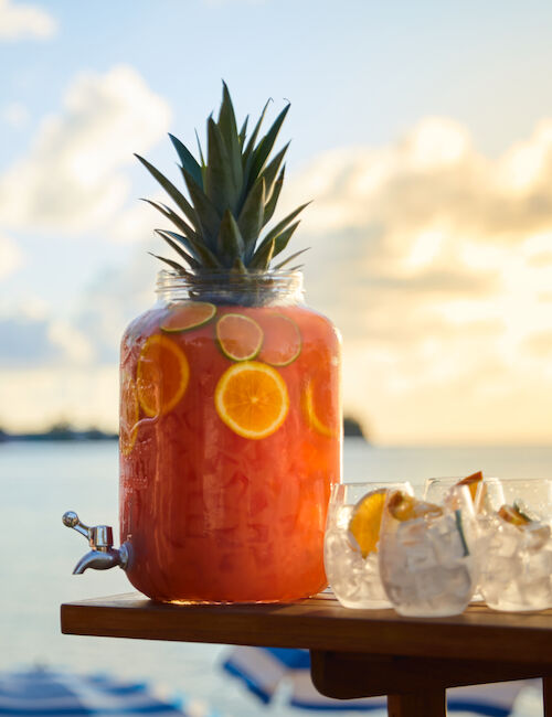 A tropical beverage jar with a pineapple top and slices of citrus sits on a table next to two glasses, set against a beachside sunset.