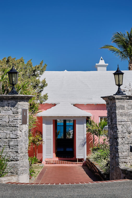 Entrance to a building with two stone pillars, a white roof, and a light pink facade surrounded by trees and plants under a clear blue sky.