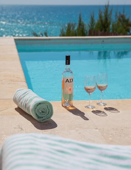 A serene poolside scene with a bottle of wine, two filled glasses, and rolled towels. The ocean and clear sky are in the background.