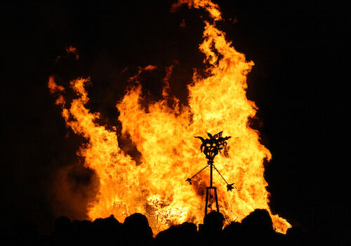 A large bonfire prominently burns in the night, with a silhouetted metal structure and crowd of shadows visible in the foreground.