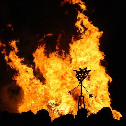 A large bonfire prominently burns in the night, with a silhouetted metal structure and crowd of shadows visible in the foreground.