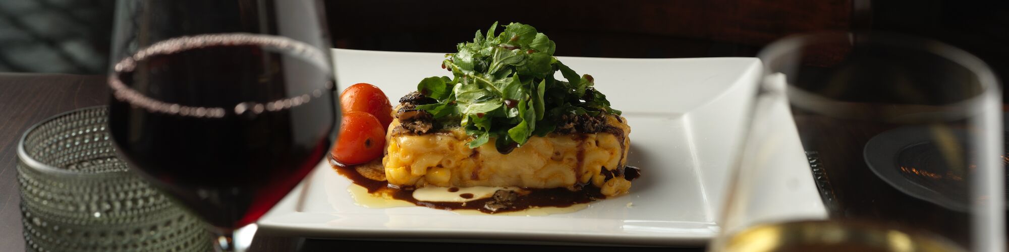 A meal is served on a white square plate garnished with greens, accompanied by two glasses of wine, one red and one white, on a table.