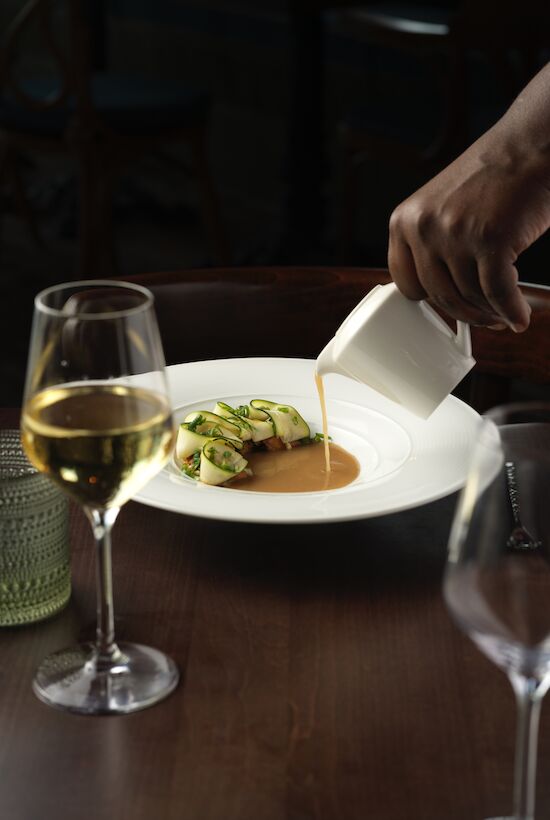 A hand pours sauce onto a plated dish with green vegetables. Two wine glasses and a green glass are on the table.