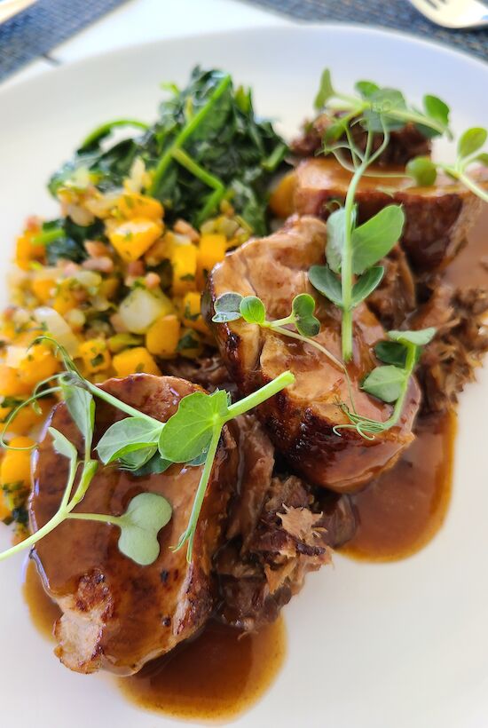 This image shows a plated dish of meat topped with microgreens, with a side of mixed vegetables and greens. The meal is served with a sauce.