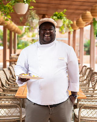 A smiling chef in a white uniform holds a dish, standing in an outdoor dining area with wooden tables and chairs surrounded by hanging plants.