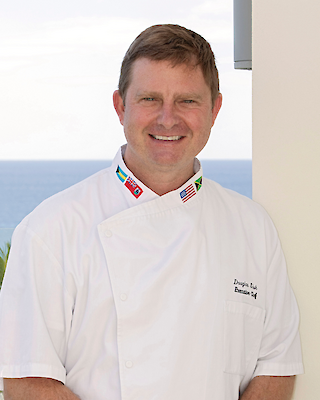 A man in a chef's uniform is standing in front of a scenic ocean view, smiling. The name on his uniform is partially visible against the white fabric.