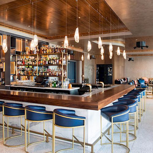 The image shows a modern, stylish bar with a long counter, blue barstools, hanging lights, and a fully stocked bar in the background.