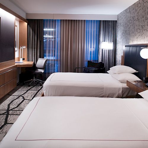 The image shows a modern hotel room with two neatly made beds, a TV, a desk with a chair, and contemporary decor and lighting.