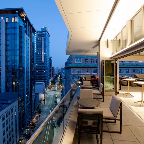 The image shows a modern city skyline with tall buildings on a busy street, viewed from a brightly lit, furnished balcony in the evening.