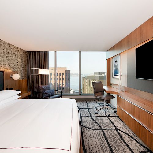 A modern hotel room with a large bed, wall-mounted TV, chair, desk, and a window offering a city and water view.