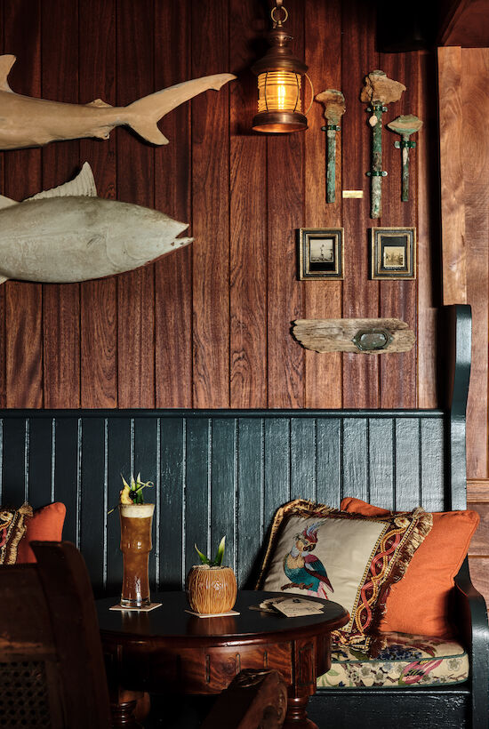 A cozy wooden interior with decorative fish, framed photos, cushions, and drinks on a table. A lantern adds a warm touch.