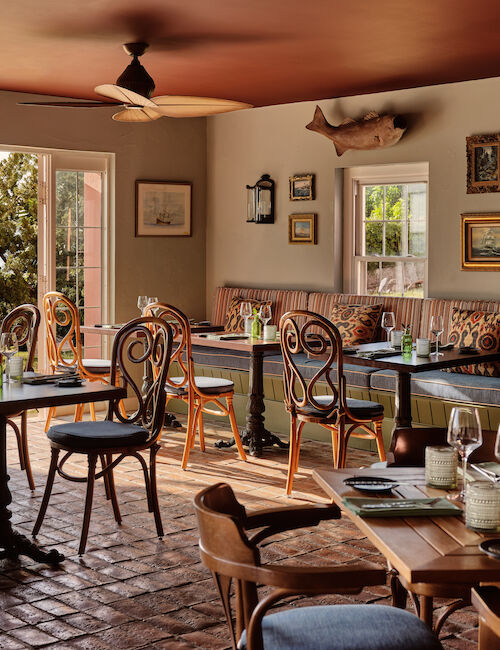 A cozy restaurant with a nautical theme, featuring wooden furniture, table settings, and wall decorations, overlooking water through large windows.