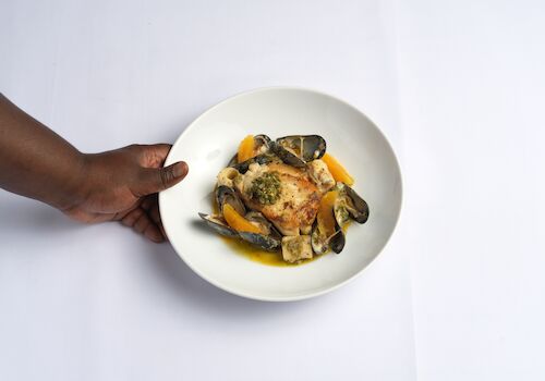 A hand holding a white plate with seafood, chicken, and vegetables in a sauce, creating a colorful dish.