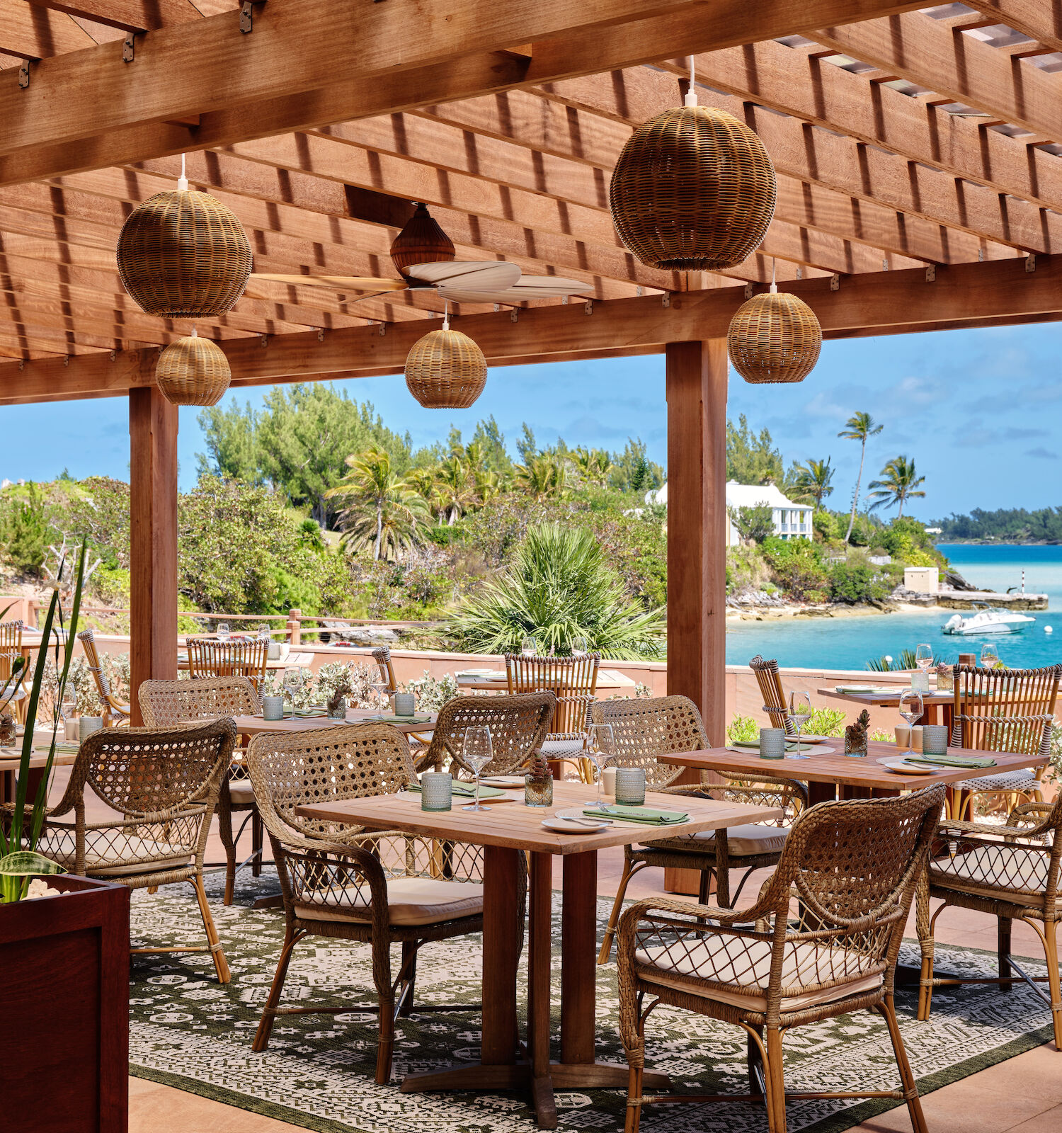 An outdoor dining area by the water, featuring wooden furniture, woven pendant lights, and a view of lush greenery and a clear sky.