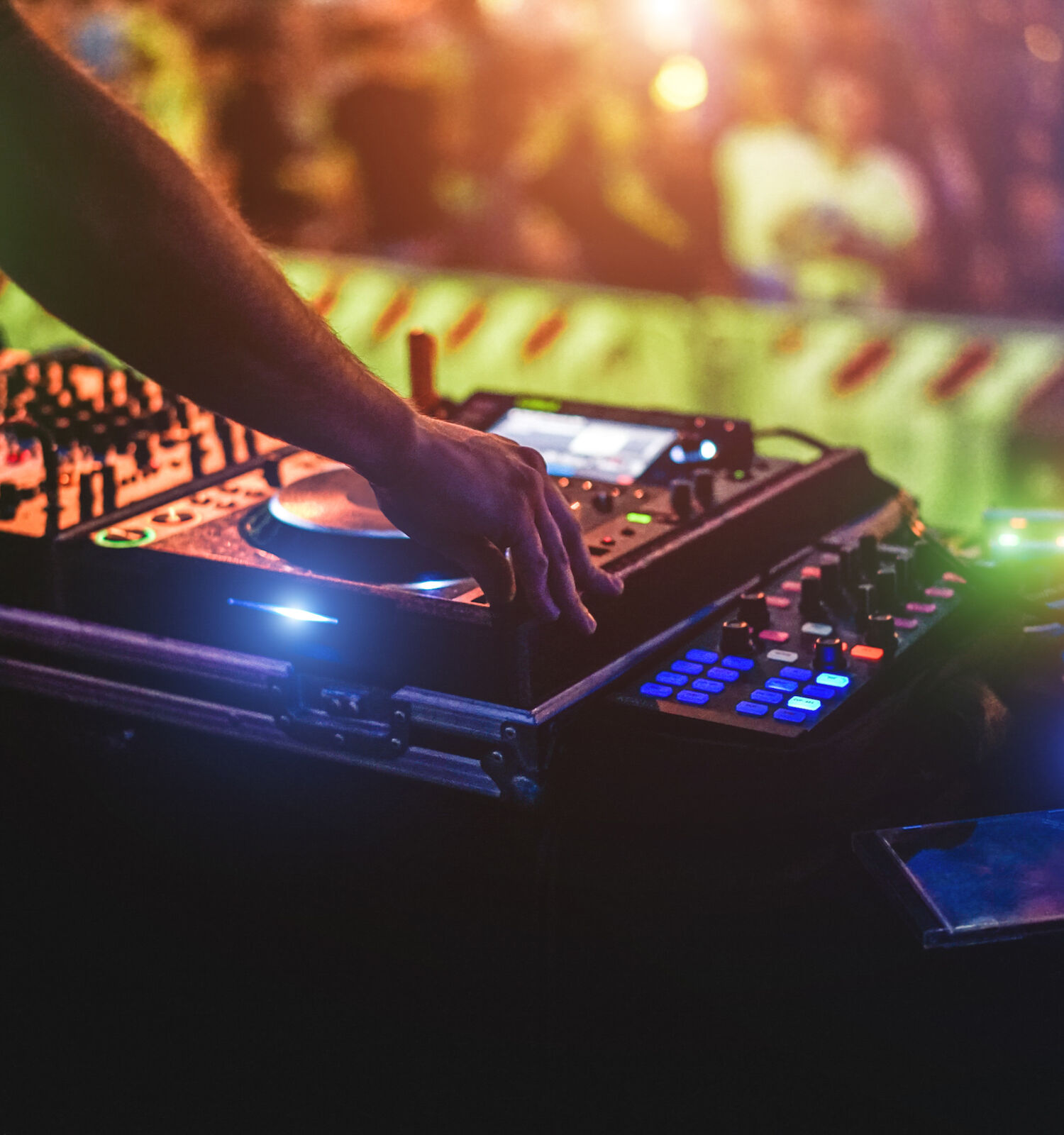 A DJ is mixing music using turntables and a laptop in a lively, colorful setting, with a blurred crowd in the background.