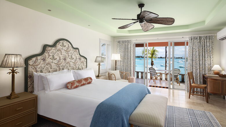 A cozy bedroom with a sea view features a bed, ceiling fan, lamps, and a desk. The balcony offers seating with ocean scenery beyond the glass doors.