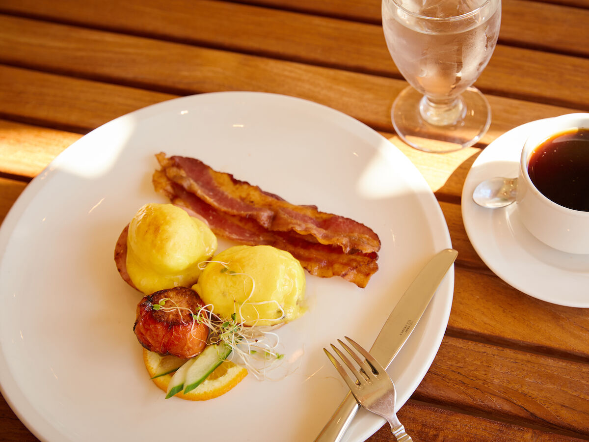 A breakfast plate with eggs benedict, bacon, vegetables, with a cup of coffee and a glass of water on a wooden table.