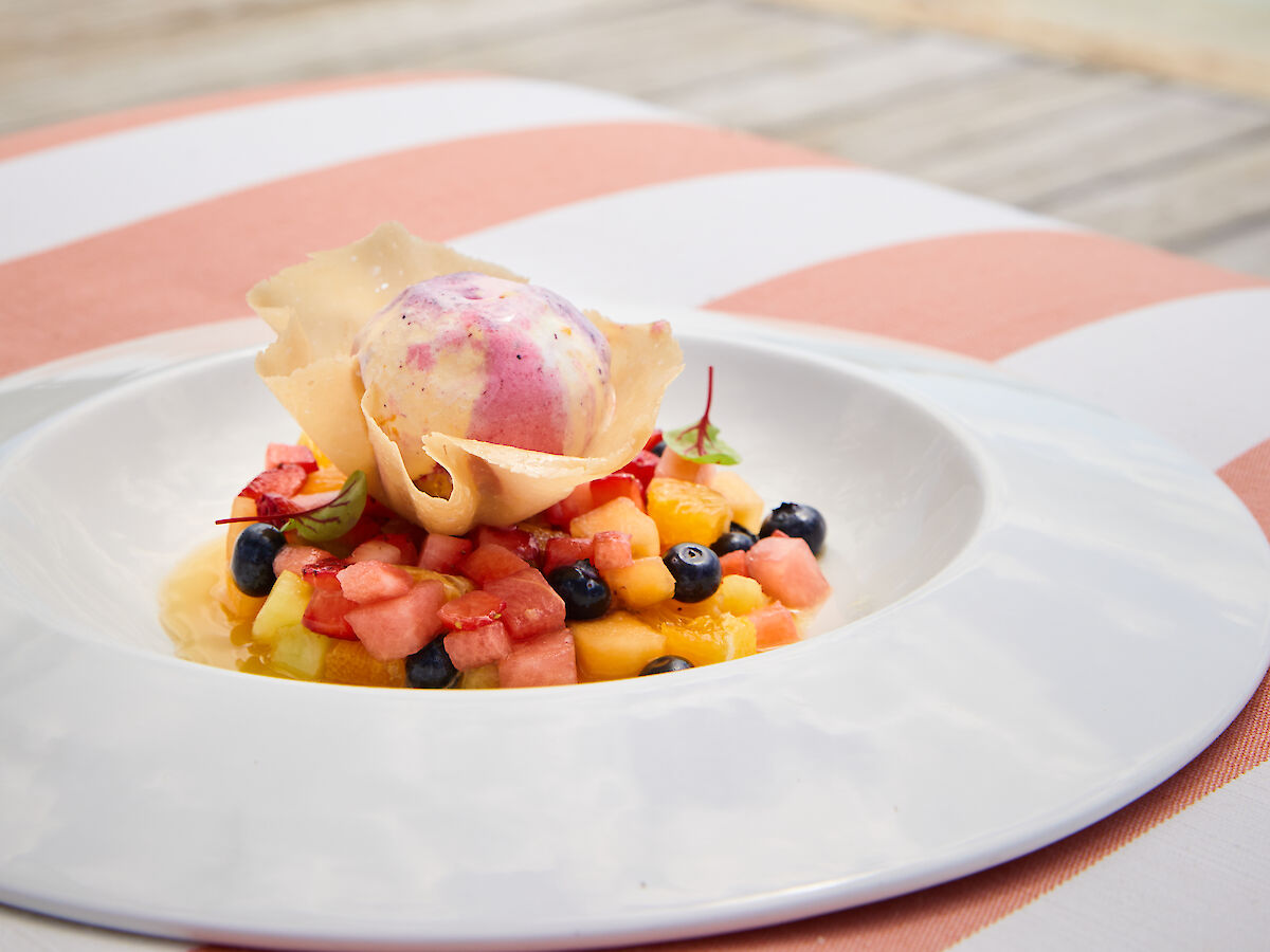 A plate of colorful fruit salad with a scoop of ice cream by a lake, set on a striped tablecloth, with trees and sky in the background.