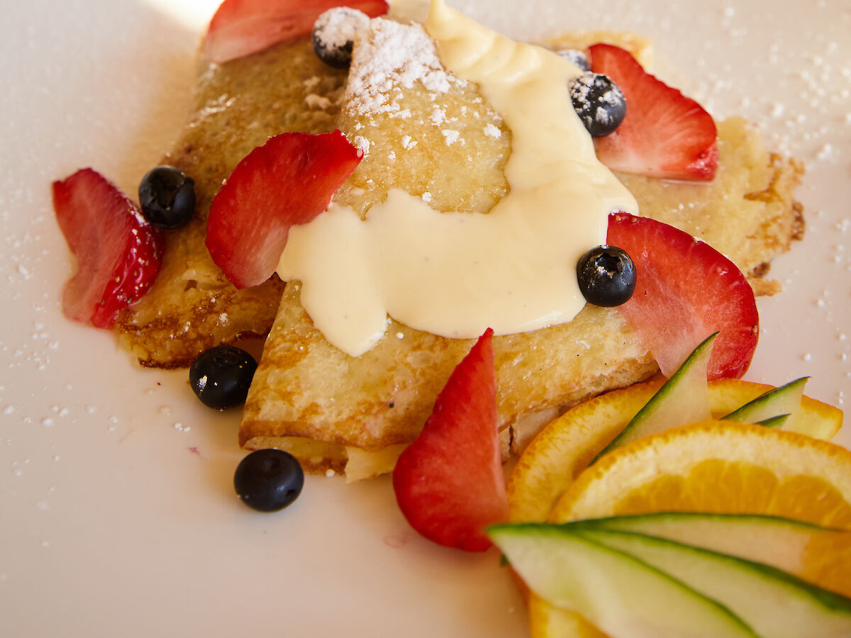 A person is pouring syrup over pancakes topped with strawberries, blueberries, and a sprinkle of powdered sugar on a plate.