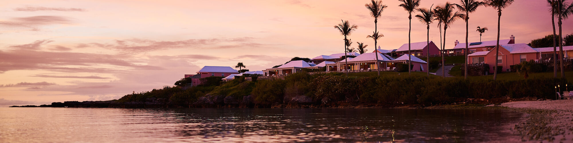 A serene beach scene at sunset with palm trees and buildings, reflecting colorful skies on the water's surface.