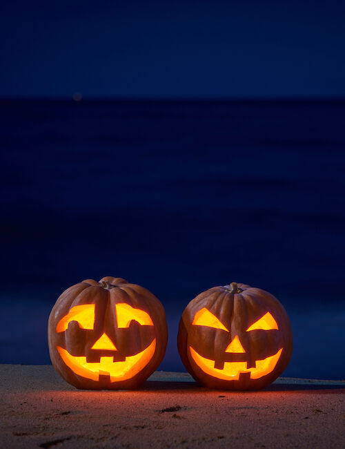 Two carved pumpkins with glowing faces sit on a beach at night, against a backdrop of dark ocean and sky.