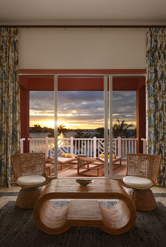 A cozy room features a wooden table, chairs, and a TV, with a balcony view of a sunset sky through large windows.