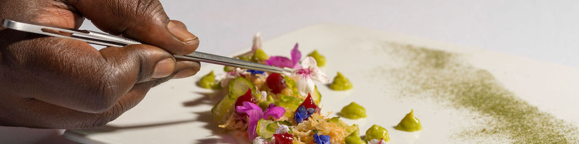 A person is arranging a colorful garnish on a white rectangular plate with a utensil, creating an artistic presentation.