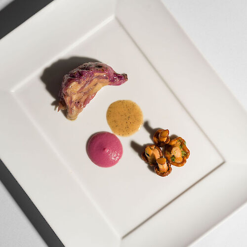 A gourmet dish with meat, two sauces, and a garnish, artfully arranged on a square white plate.