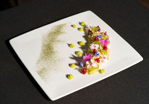 A gourmet dish with colorful flowers, avocado dollops, and a green powder on a square plate.