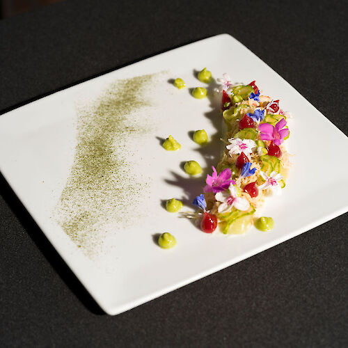 A gourmet dish with colorful flowers, avocado dollops, and a green powder on a square plate.