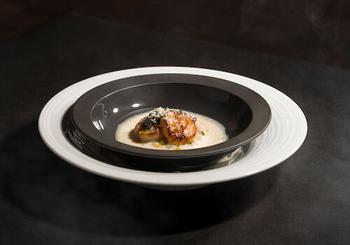 A gourmet dish with a seared piece of meat or seafood atop a creamy sauce, served in a deep black bowl on a white plate.