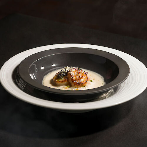 A gourmet dish with a seared piece of meat or seafood atop a creamy sauce, served in a deep black bowl on a white plate.