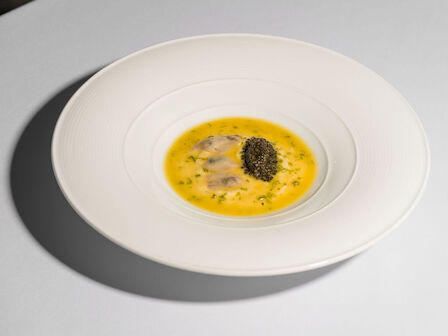 A white plate with a creamy yellow soup, garnished with herbs and topped with what appears to be caviar or a similar ingredient.