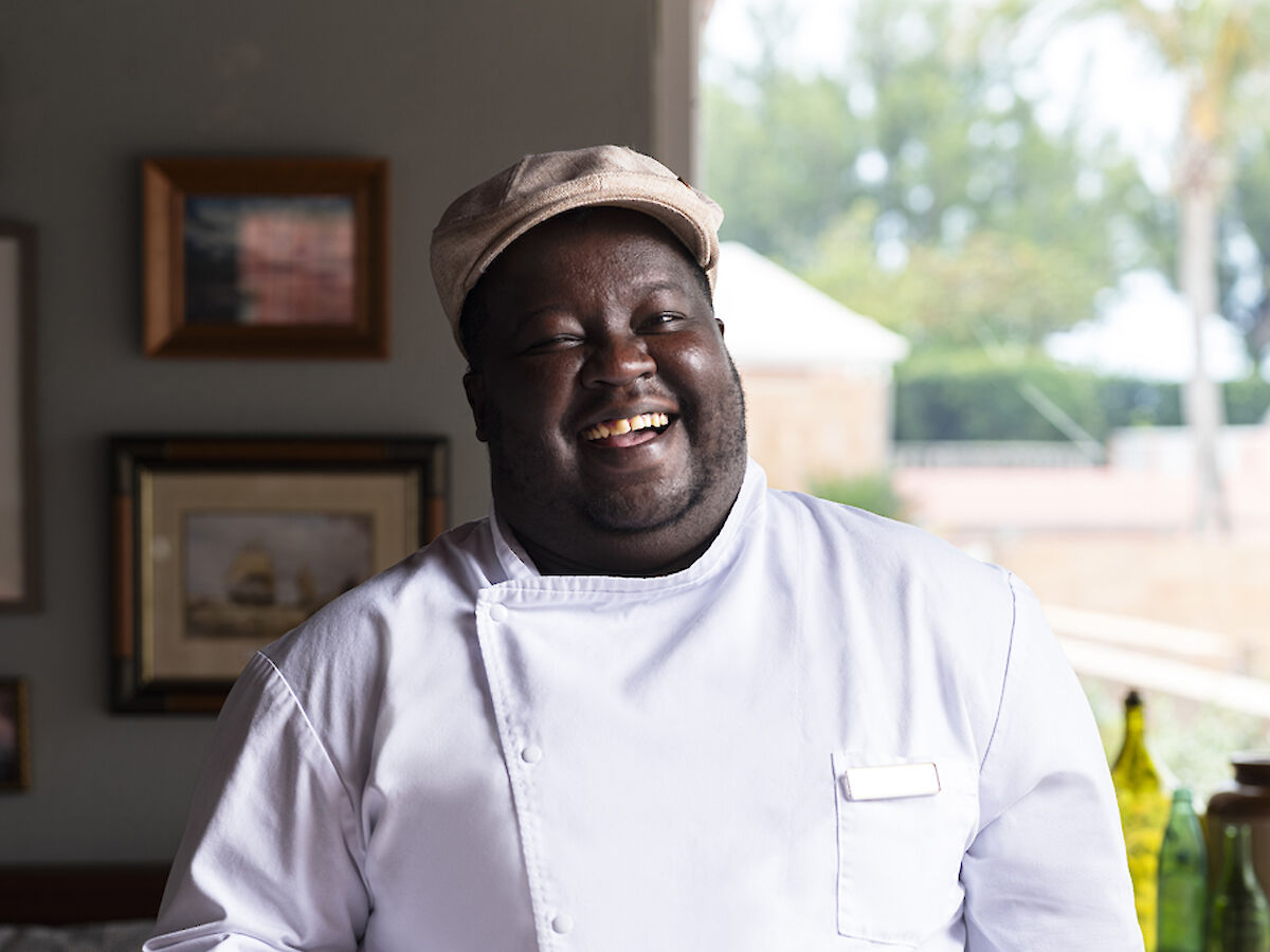 A chef in a white uniform holds a plate with food, standing in a warmly decorated dining area, smiling and looking content.