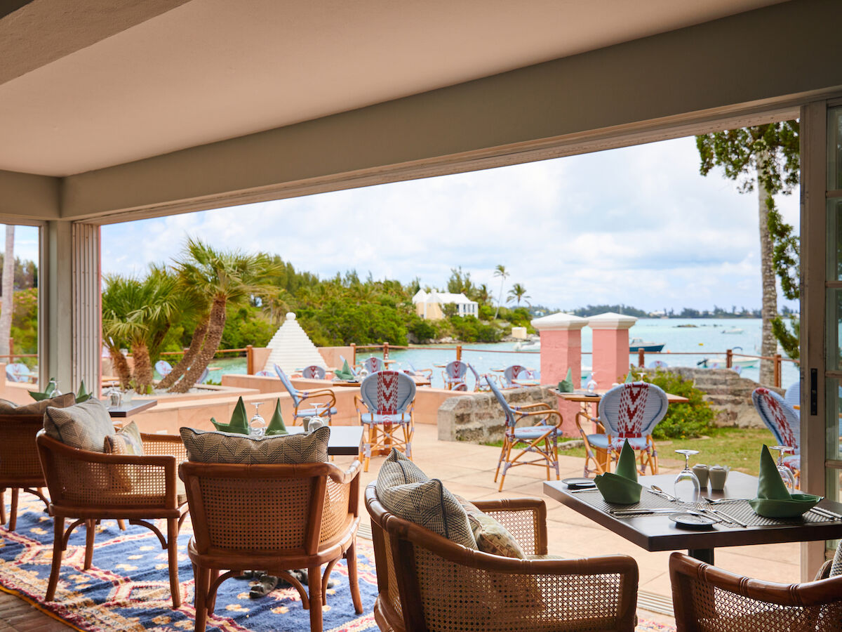 A seaside terrace with wicker chairs overlooking the ocean, surrounded by palm trees and decorative pink walls on a sunny day.