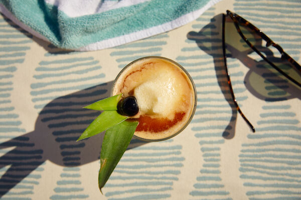 A tropical drink garnished with pineapple leaves and a cherry sits on a patterned surface beside sunglasses and a towel.